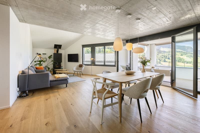 Modern living and dining area with wooden flooring, large windows, and a scenic view. Features a grey sofa, dining table with chairs, pendant lights, and decorative items.