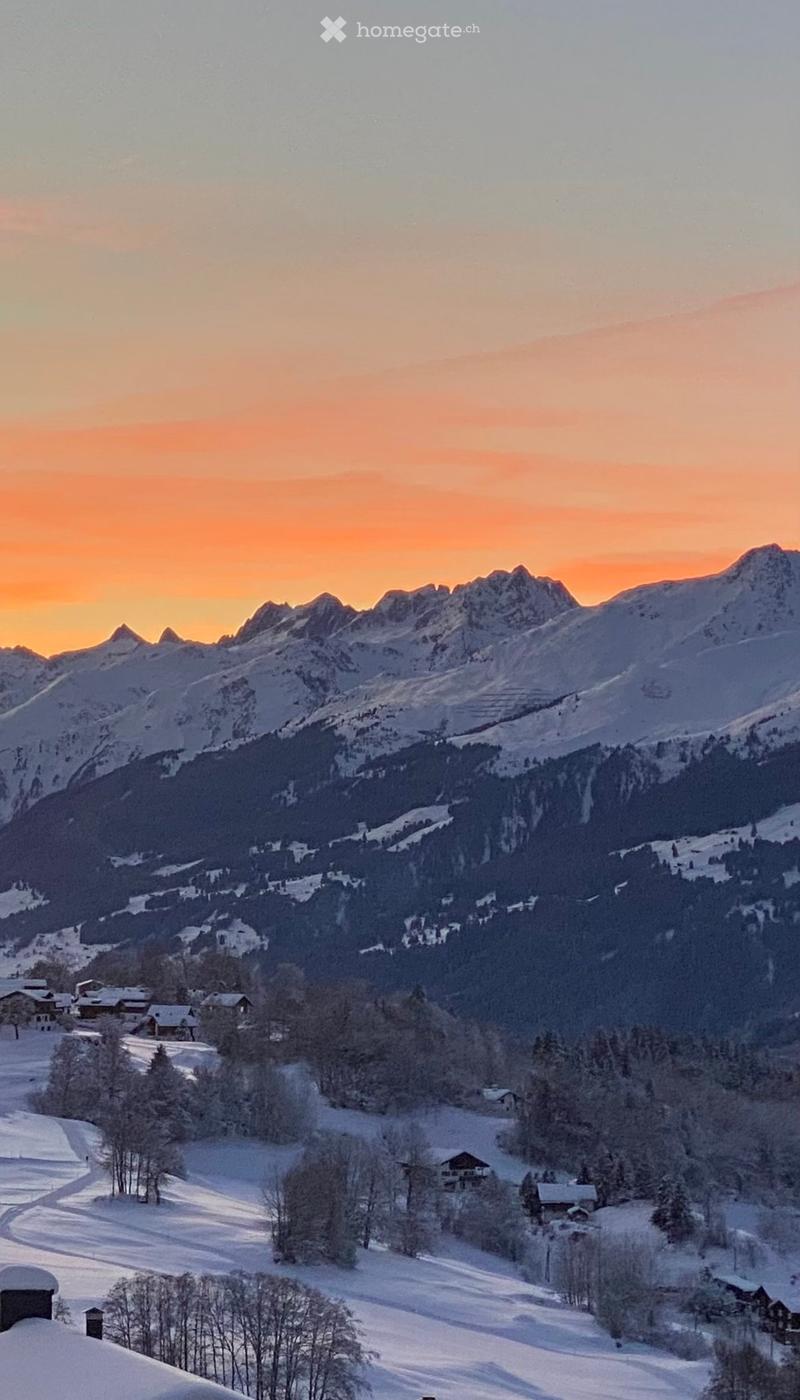 A serene mountain landscape at sunset, featuring snow-covered peaks, a forested valley, and scattered houses nestled among trees.