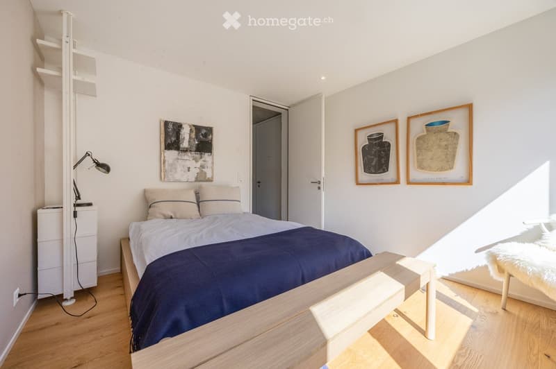 Modern bedroom with a wooden bed, nightstand, lamp, and framed artwork on the walls. The room features light-colored walls and wooden flooring.