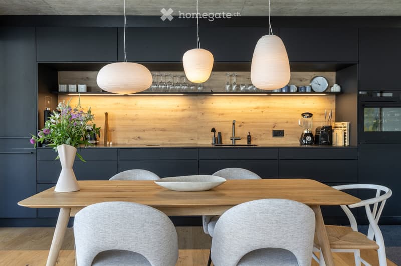 Modern kitchen with dark grey cabinets, wooden countertops, and pendant lights. Includes a dining table with chairs, a vase with flowers, and various kitchen appliances.