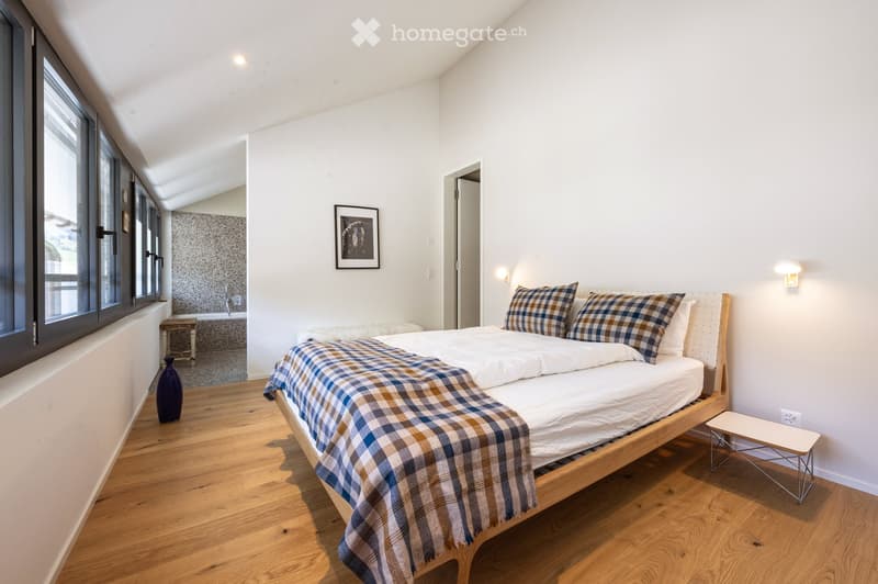 Bright bedroom with wooden flooring, a large bed with plaid bedding, a small stool, a vase, and a picture frame on the wall. A small seating area with a stool and a bathtub is visible through an open doorway.