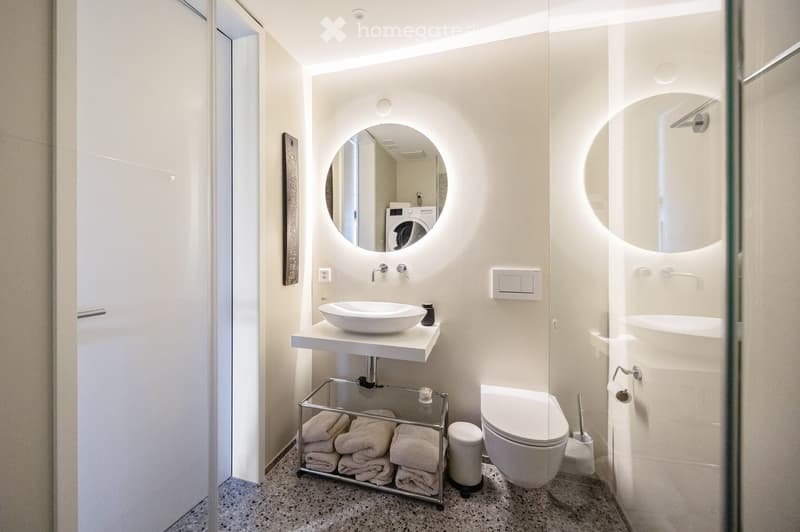 Modern bathroom with white walls, a floating sink, a round mirror, a toilet, and a towel rack holding neatly folded towels.