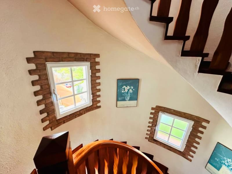 A view from above of a hallway with wooden railings, two windows with decorative wooden frames, and two framed artworks on the wall.