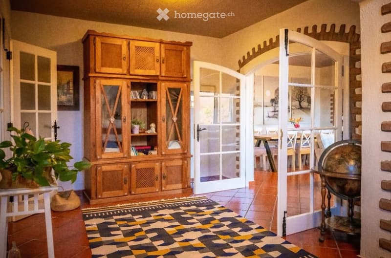 A well-lit hallway with a wooden cabinet, a patterned rug, and an open doorway leading to a dining area with a table and chairs.