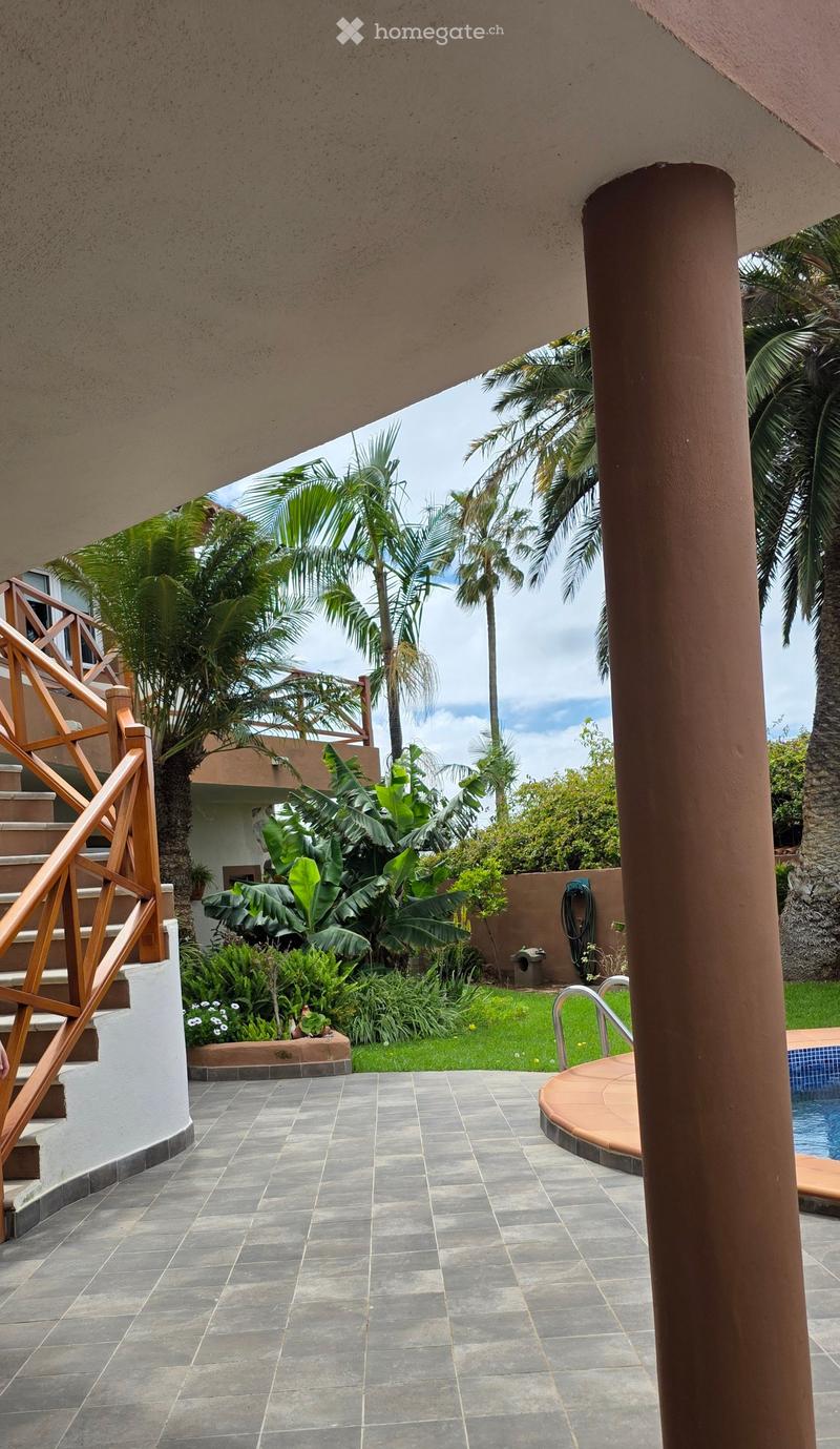 Outdoor patio with tiled flooring, wooden staircase, lush greenery, and a swimming pool.