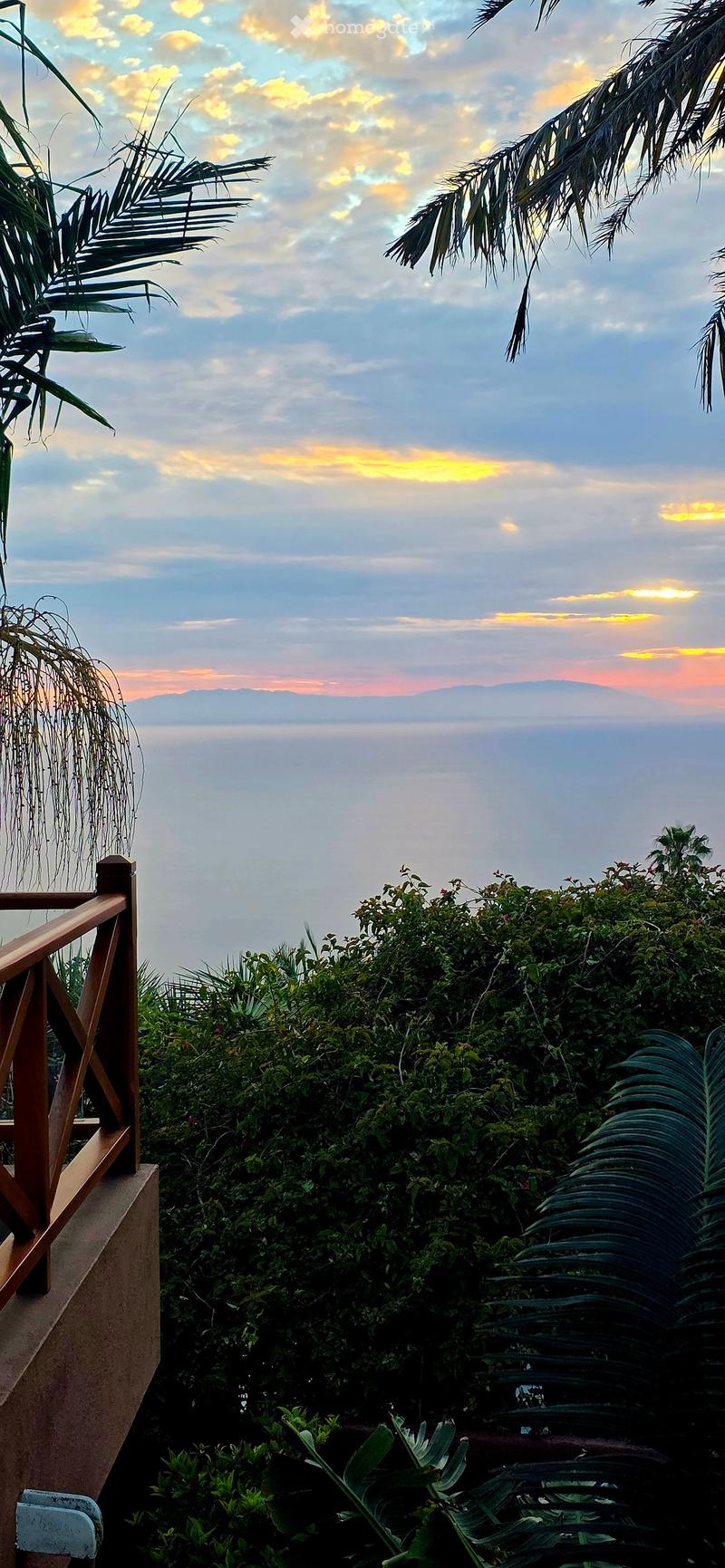 Balcony with wooden railing, lush greenery, and a scenic view of the ocean and mountains at sunset.