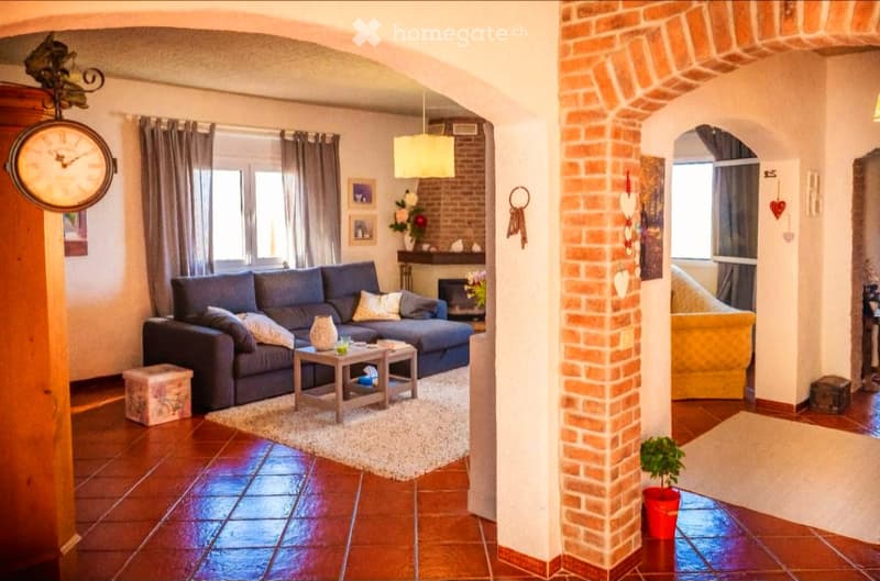 Living room with a blue sectional sofa, coffee table, fireplace, decorative clock, and potted plant. The space features exposed brick walls and large windows with curtains.