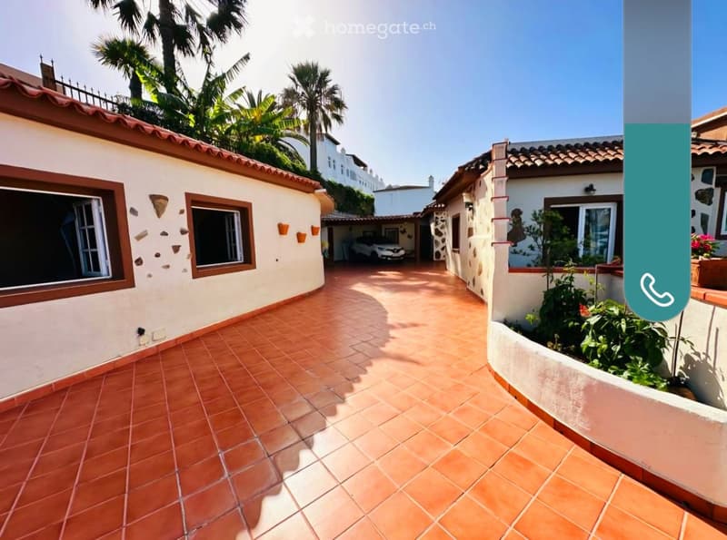 Courtyard with terracotta tiles, white walls, decorative stone accents, palm trees, and a parked car in the background.