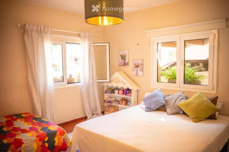 Cozy bedroom with a double bed, colorful bedspread, pillows, a small nightstand with a mirror, and a decorative shelf with books and items. Large windows with white curtains and a hanging lamp provide natural light.