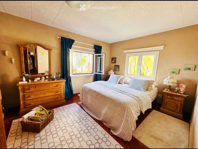 Cozy bedroom with a large bed, nightstand, dresser, mirror, and decorative items. The room features wooden flooring, large windows with blue curtains, and a patterned rug.