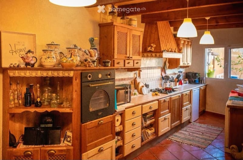 The kitchen features wooden cabinets, a stainless steel oven, microwave, and countertop. There are decorative items on the shelves, including teapots and glassware. The space is well-lit with hanging lights and has a cozy, rustic design with terracotta ti
