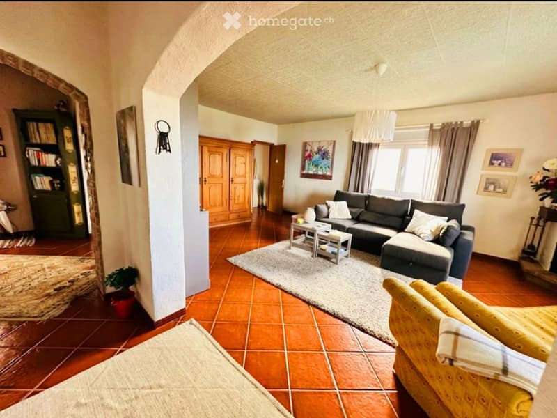 Living room with terracotta tile flooring, a grey sectional sofa, a yellow armchair, a coffee table, a bookshelf, and a large window with curtains.