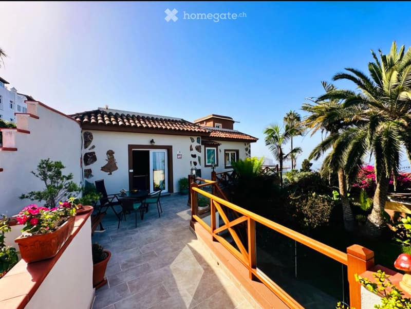 Balcony with tiled floor, wooden railing, outdoor furniture, potted plants, and a view of a house and garden with palm trees.
