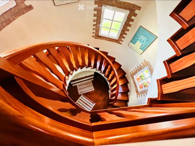 Spiral wooden staircase with a wooden handrail, light-colored walls, and a small window at the top.