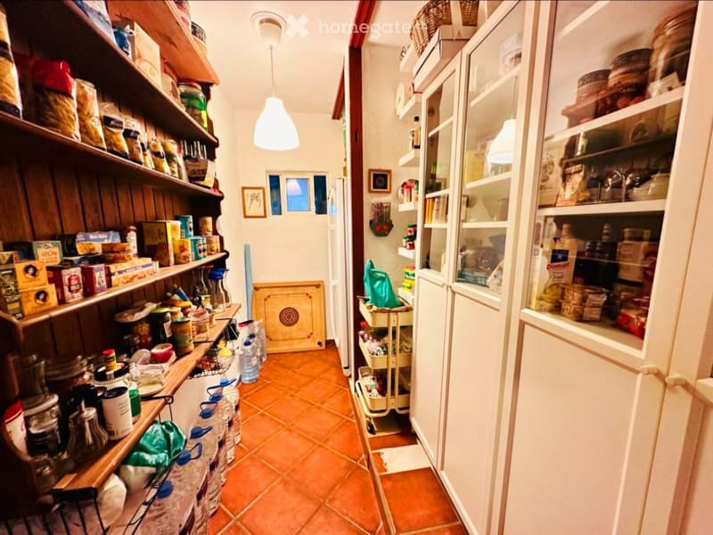 A cozy kitchen with wooden shelves filled with jars, cans, and pantry items, a refrigerator, and a small kitchen cart with additional supplies.