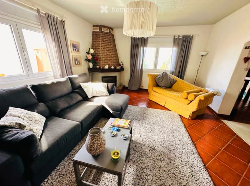 Living room with a dark grey sectional sofa, a yellow armchair, a coffee table with a vase and candle, fireplace with picture frames, and large windows with curtains.