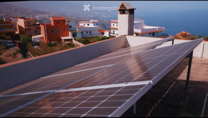 A rooftop with solar panels installed, overlooking a coastal town with houses and a view of the sea and mountains.