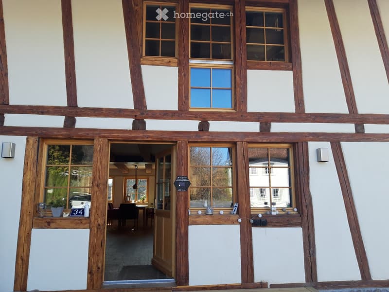 Traditional wooden-framed building with white plaster walls, large windows, and a visible interior dining area with tables and chairs.