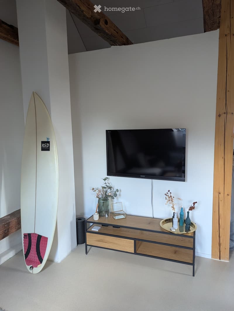Modern living room with a flat-screen TV mounted on the wall, a wooden TV stand with drawers, decorative vases, and a surfboard leaning against the wall.