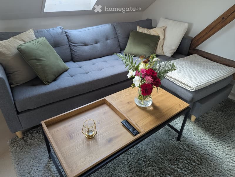 Modern living room with a grey sectional sofa, wooden coffee table, decorative vase with flowers, and a candle holder.