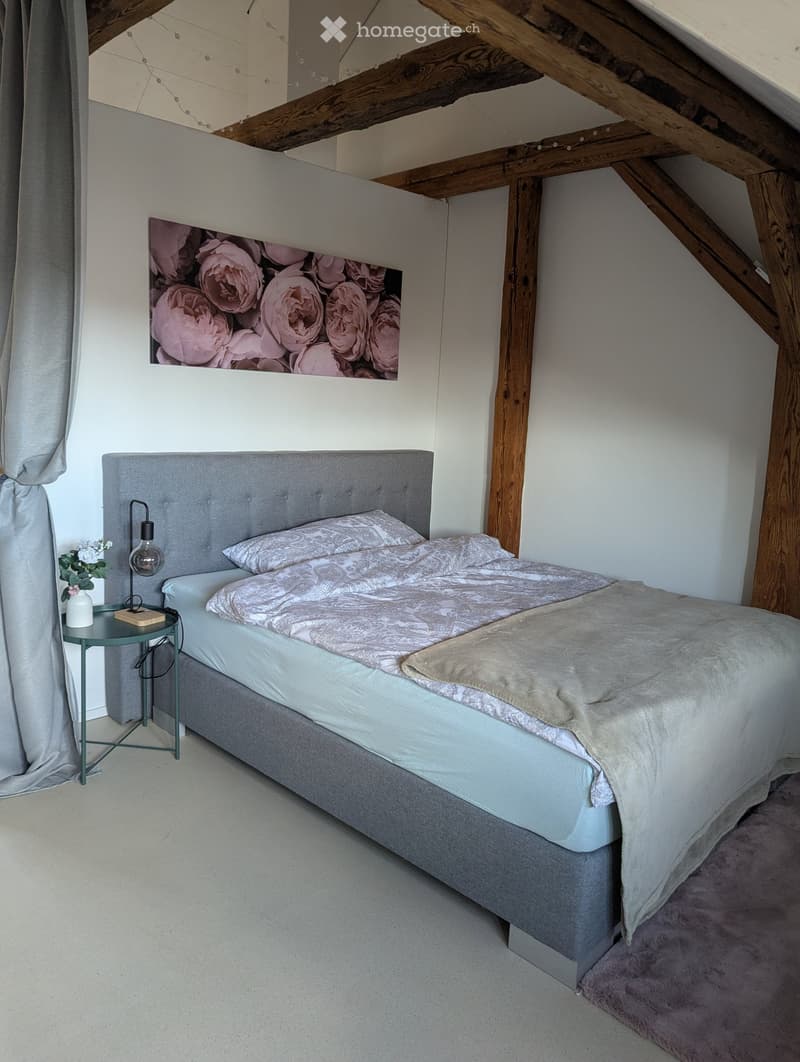 Cozy attic bedroom with wooden beams, a grey upholstered bed, decorative pillows, a nightstand with a lamp, and a vase with flowers. A floral picture adorns the wall.