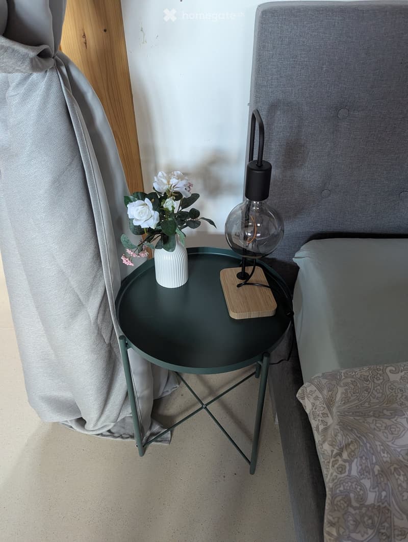 A cozy bedroom corner with a grey sofa, a round dark green side table, a white vase with white and pink flowers, and a modern black lamp with a glass shade.