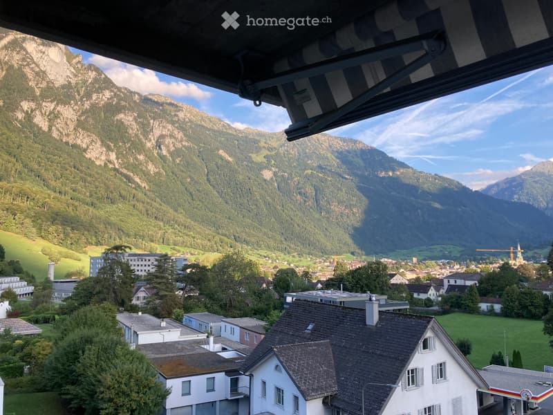 Balcony view overlooking a town nestled in a valley surrounded by mountains, with houses, trees, and a clear blue sky.