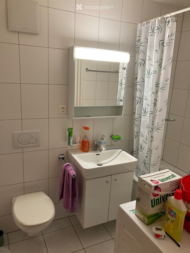 Modern bathroom with a white sink, mirror with built-in lighting, toilet, towel, and cleaning supplies on a shelf.