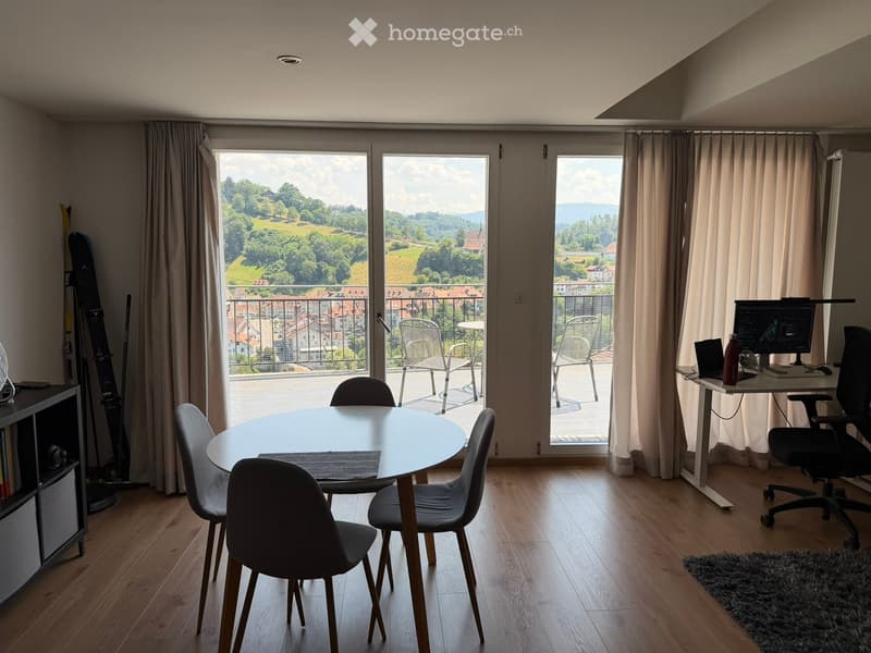 Bright room with wooden flooring, large windows, and a round dining table with chairs. A desk with a laptop and office chair is visible, along with a balcony overlooking a scenic landscape.