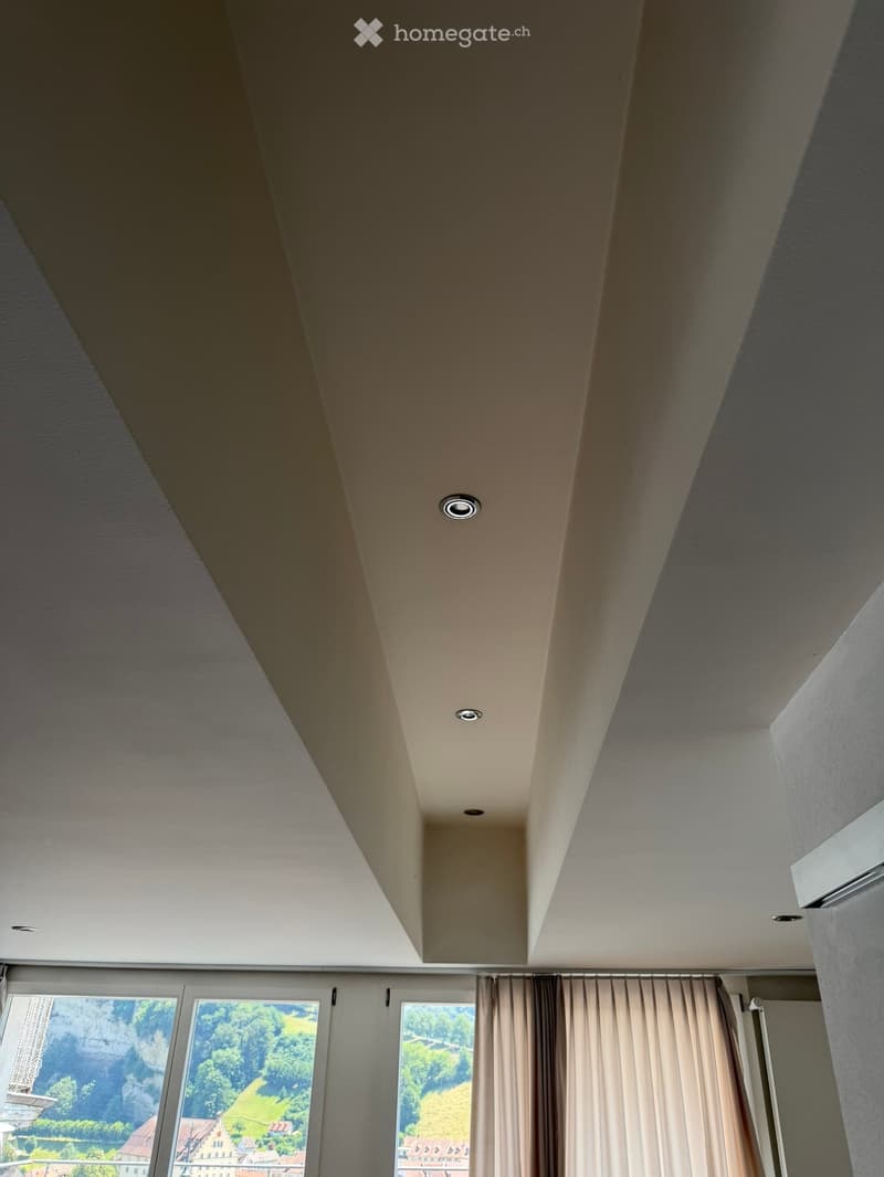 Empty room with large windows, beige curtains, and recessed ceiling lights.