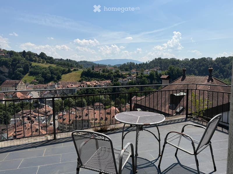 Balcony with a metal table and two chairs, overlooking a town with red-roofed houses and green hills.