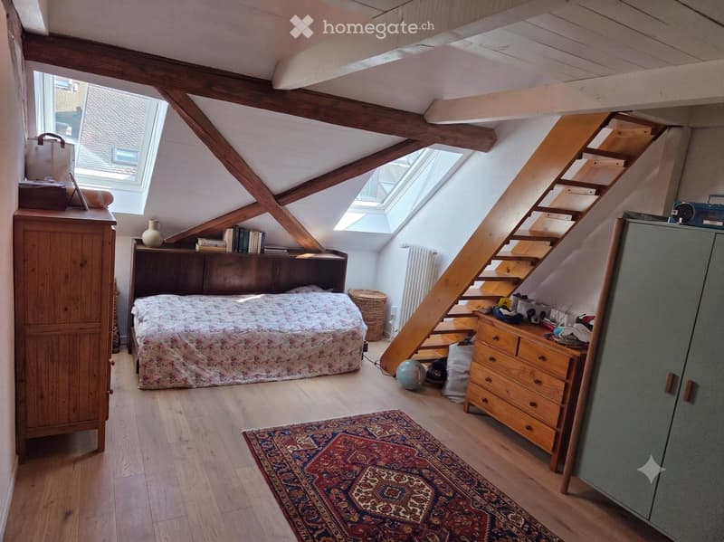Cozy attic bedroom with wooden beams, skylights, and a staircase leading to another level. Features a bed with a floral duvet, a wooden wardrobe, a bookshelf, a drawer unit, and a patterned rug.