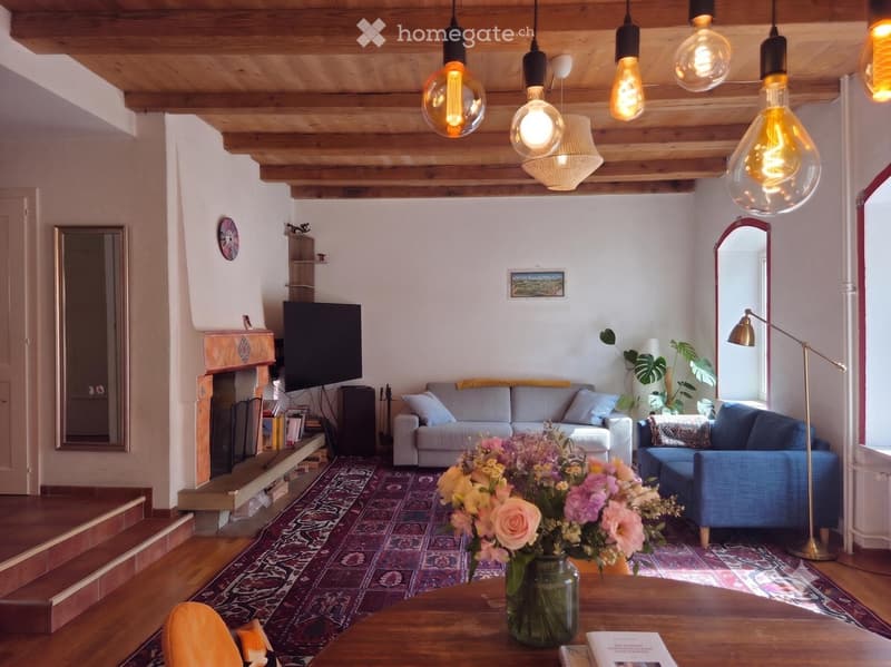 Cozy living room with exposed wooden beams, a fireplace, a sofa, a chair, a coffee table with a flower vase, and pendant lights.