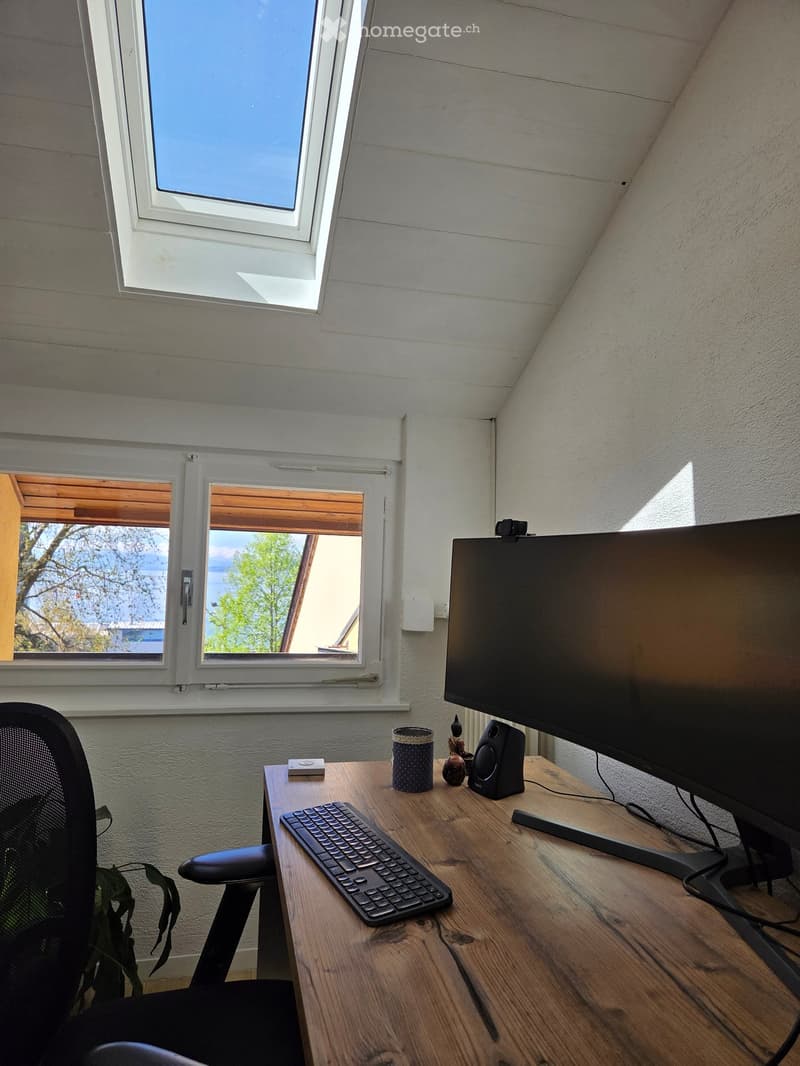 A small, well-lit room with a wooden desk, a black office chair, a large monitor, a keyboard, a speaker, a cup, and a small decorative statue. The room has white walls, a slanted ceiling, and large windows offering a view of the outdoors.