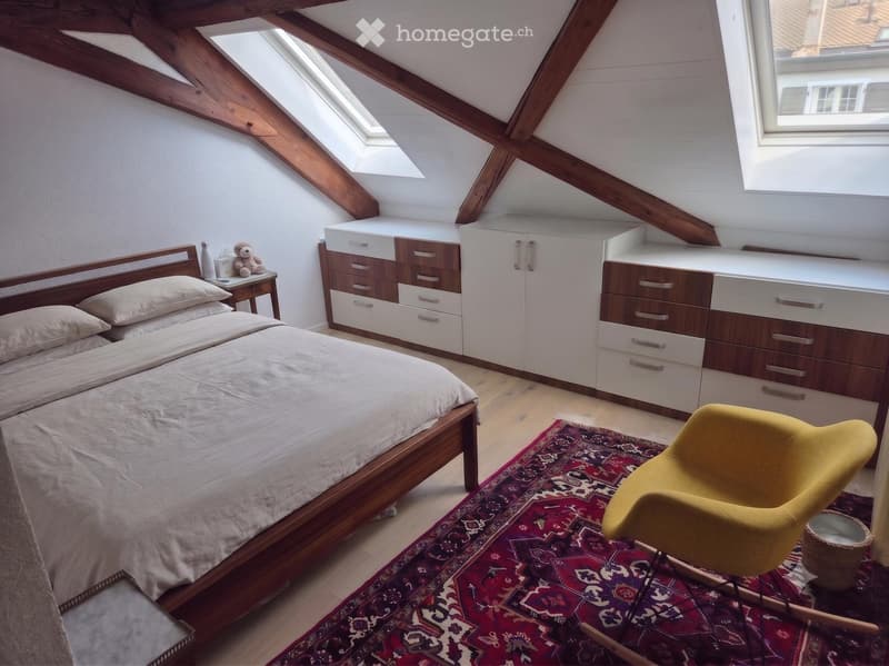 Cozy attic bedroom with wooden beams, skylights, a bed with white bedding, a nightstand with a lamp, a yellow armchair, and built-in cabinets.