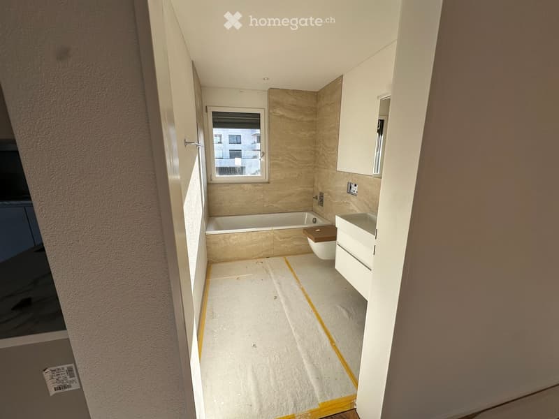Modern bathroom with a bathtub, toilet, sink, and cabinet. Light-colored tiles and a window provide natural light.