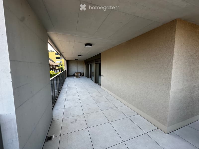 Empty balcony with tiled floor, glass railing, and concrete walls. A few boxes are placed near the entrance.