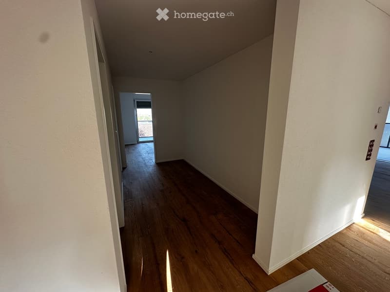 Empty hallway with wooden flooring and white walls, leading to an open doorway with natural light.