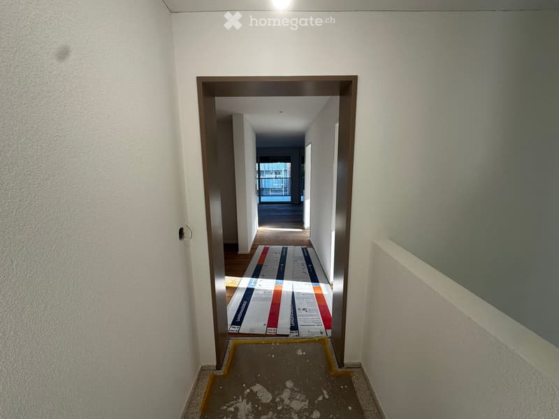 Empty hallway with white walls, a wooden floor, and a partially visible room at the end. There are some protective sheets on the floor.