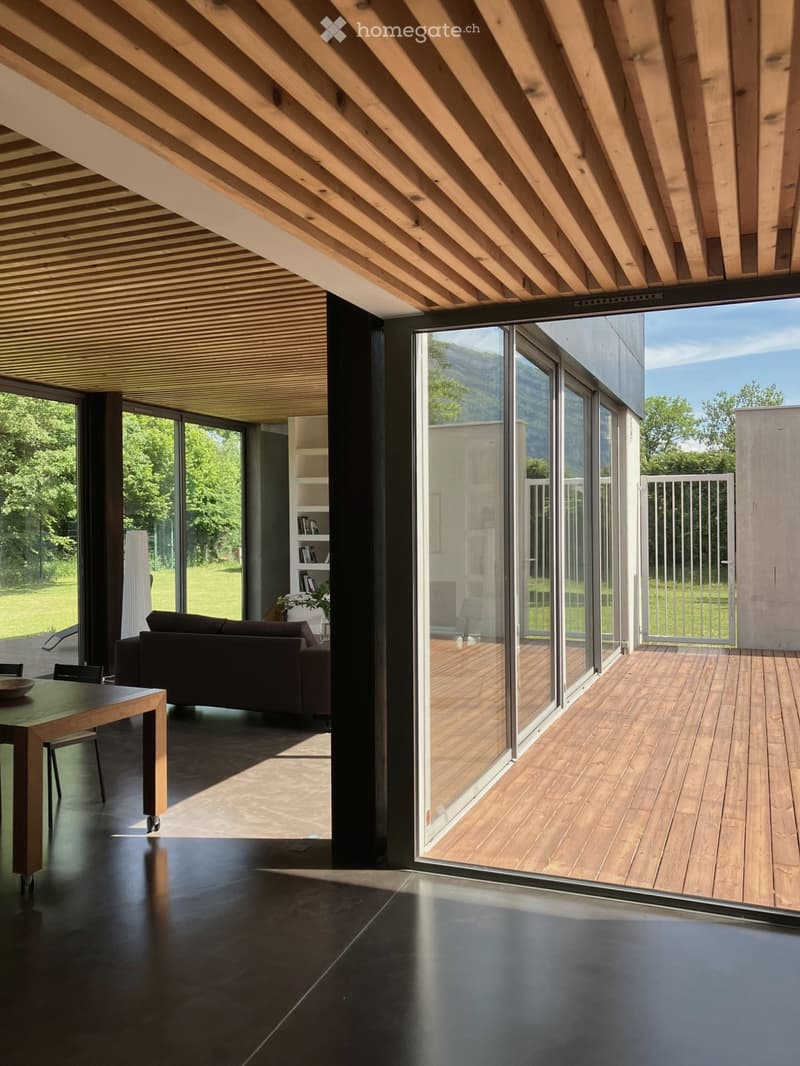 Modern living room with dark wooden flooring, large glass windows, and a view of a green landscape. Features a dark brown sofa, a wooden dining table with chairs, and a built-in shelf unit.
