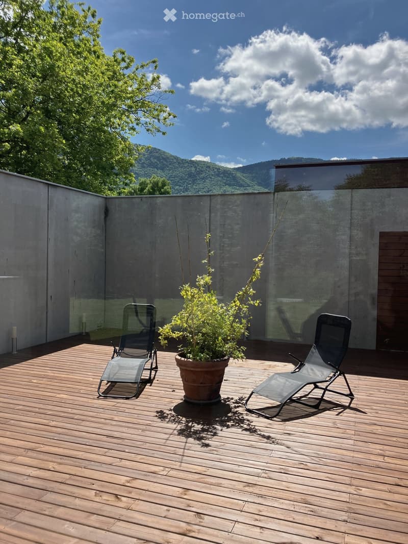 Wooden deck with two lounge chairs and a potted plant, surrounded by a low concrete wall and a glass railing, with a scenic mountain view in the background.