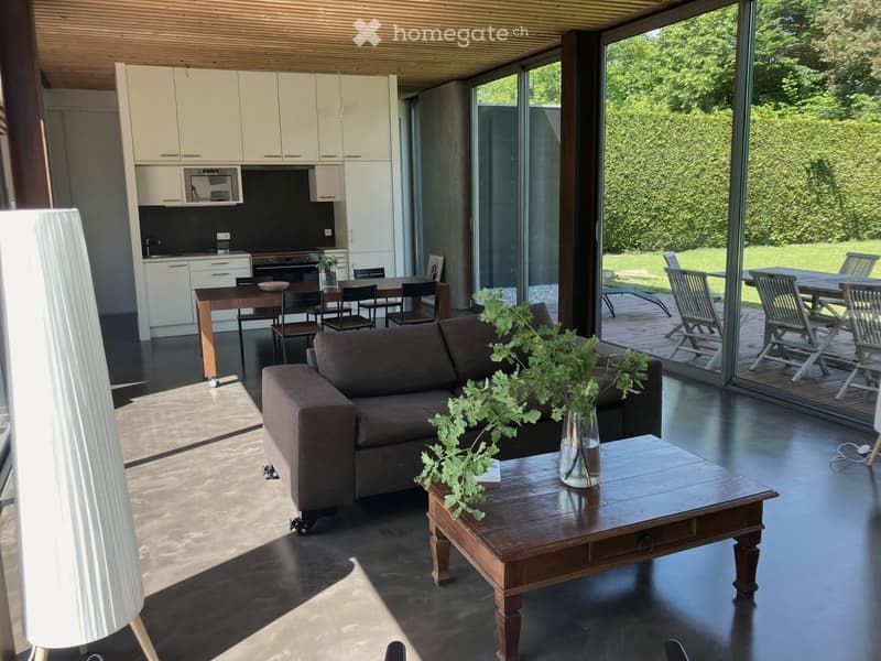 Modern living and dining area with a dark-toned sofa, coffee table, dining table with chairs, and a fully equipped kitchen with white cabinets and stainless steel appliances. Large glass windows open to a patio with outdoor furniture.