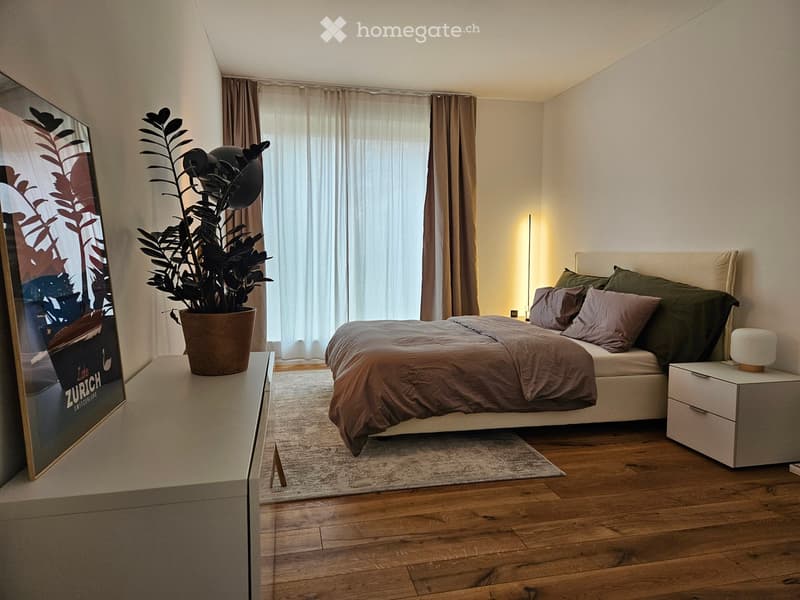 Modern bedroom with wooden flooring, a large bed with pillows and a duvet, a nightstand with a lamp, a plant on a side table, and a large picture frame on the wall.