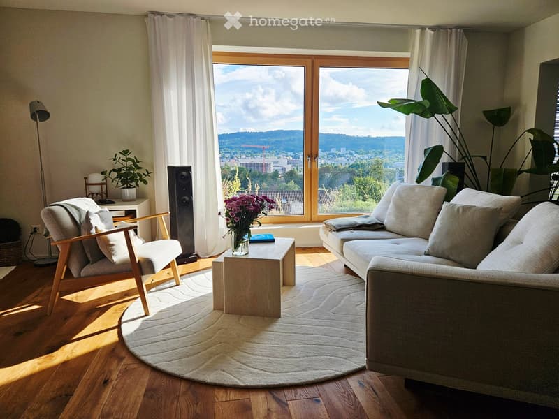 Bright living room with large windows offering a scenic view, featuring a sofa, armchair, coffee table, floor lamp, and decorative plants.