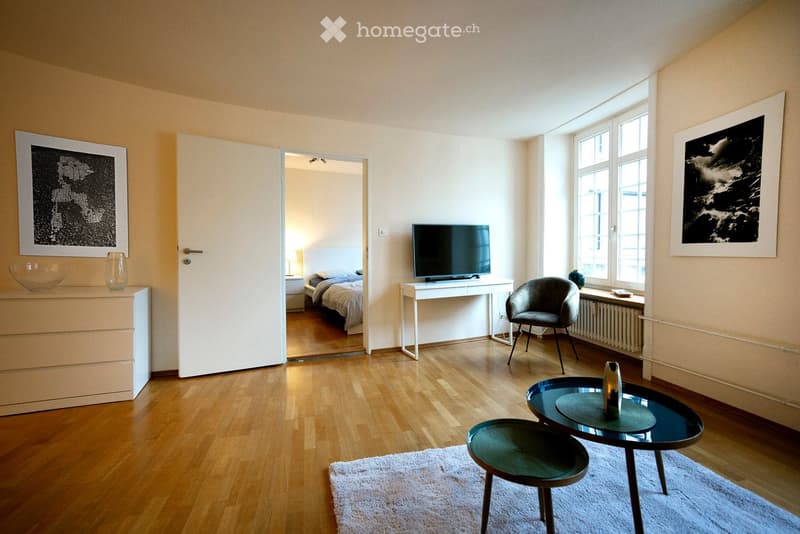 Wooden floor, beige walls, open door leading to a bedroom, TV on a stand, white side table, armchair by the window, radiator, two artworks on the wall