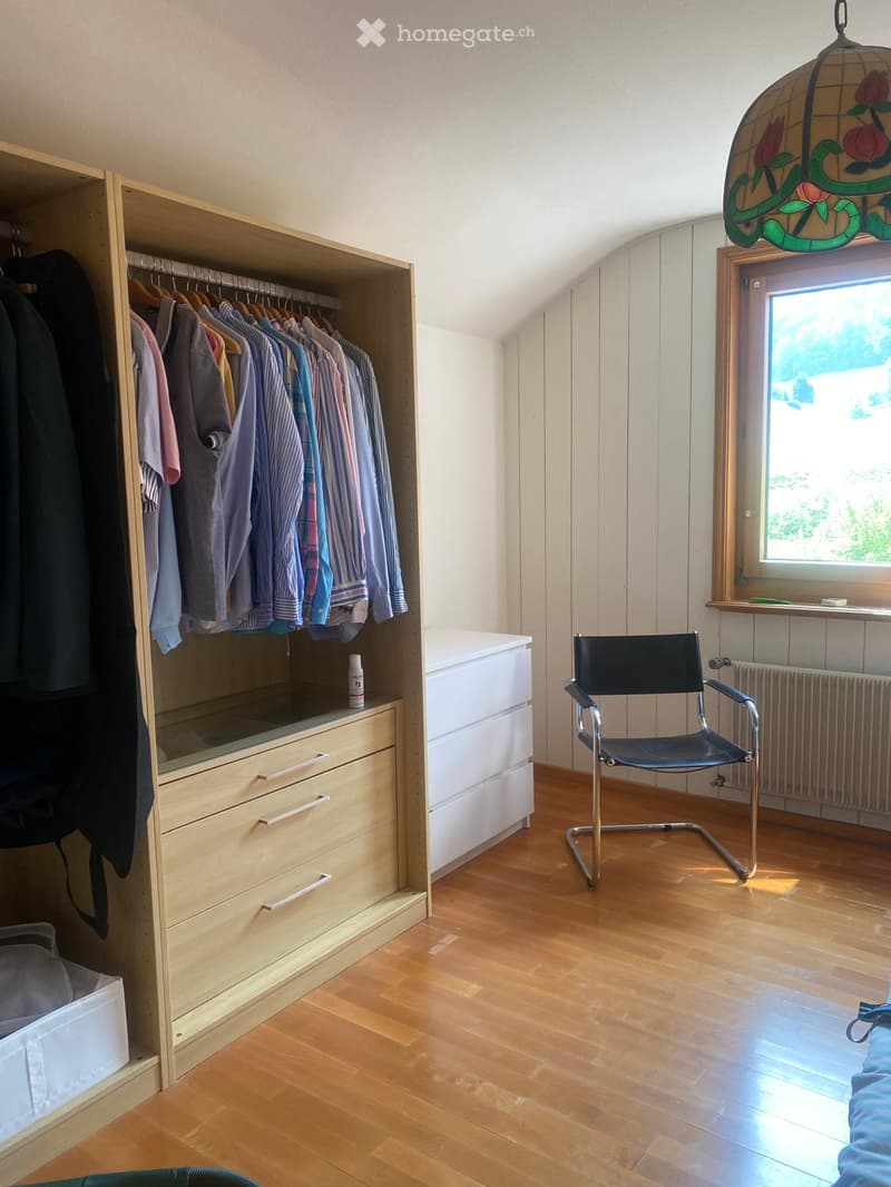A well-lit room with wooden flooring, a large built-in wardrobe with hanging clothes and drawers, a chair, a radiator, and a window with a view.