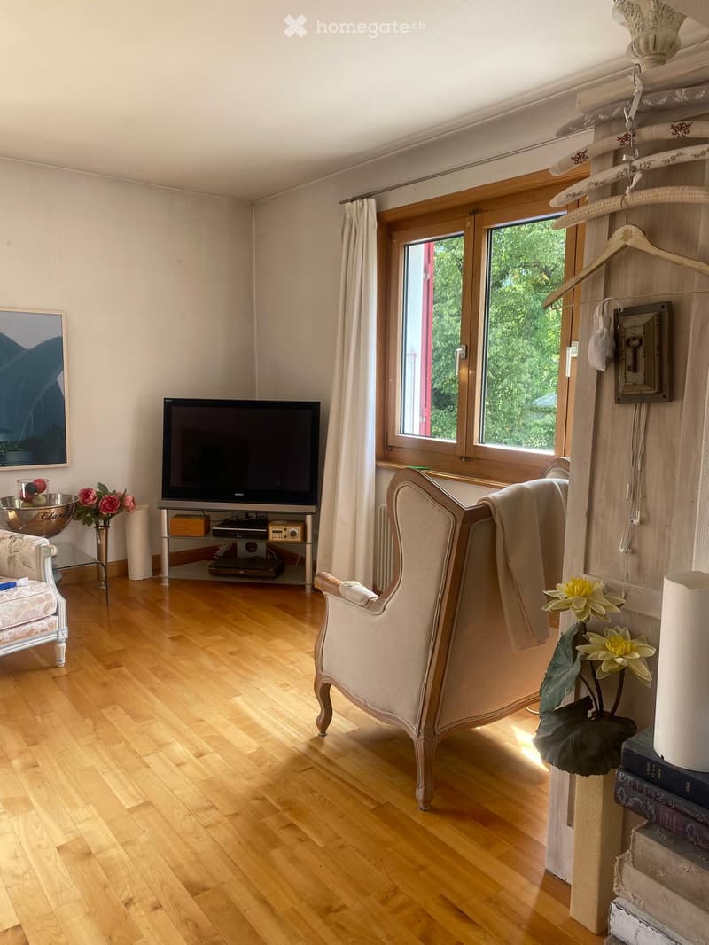 A well-lit living room with wooden flooring, a large window with curtains, a beige armchair, a white sofa, a television on a stand, a small table with a flower arrangement, and a painting on the wall.