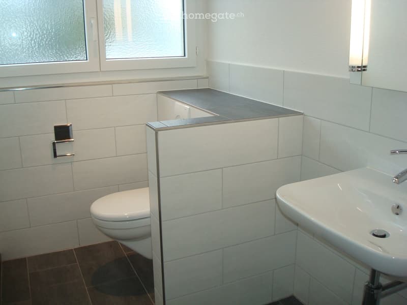 Modern bathroom with white tiles, a toilet, a sink, a mirror, and a window with frosted glass.