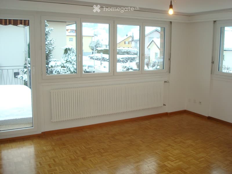 Empty room with wooden flooring, white walls, large windows, radiator, and a glass door leading to a balcony.