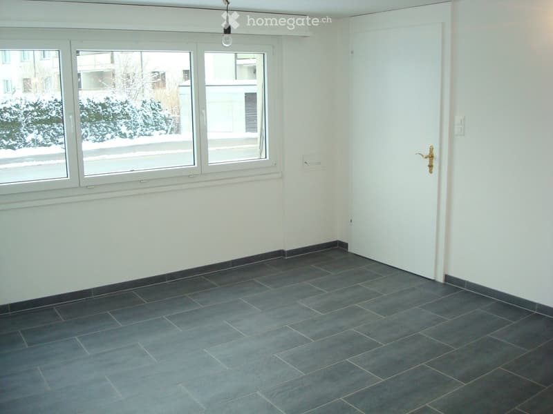 Empty room with grey tiled floor, white walls, large windows, and a door with a golden handle.